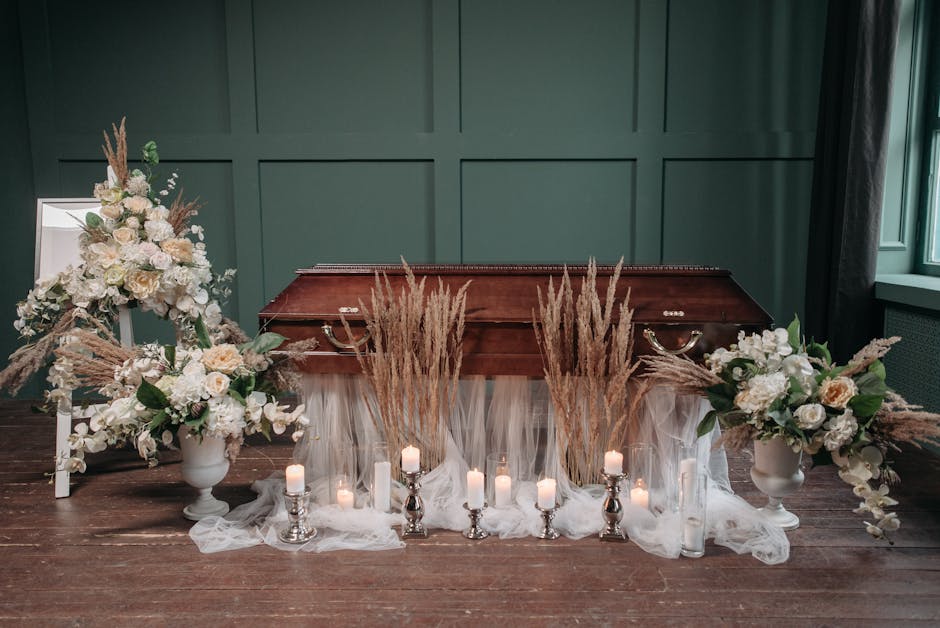 A floral arrangement set up for a funeral decoration featuring two large white urn-style vases filled with cream and white roses, peonies, and greenery, accented with pampas grass and tall wheat stalk