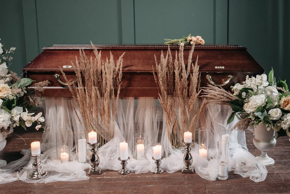A wooden casket adorned with floral arrangements featuring white and cream roses, white orchids, and greenery, surrounded by tall beige pampas grass and white pillar and tea light candles in glass and