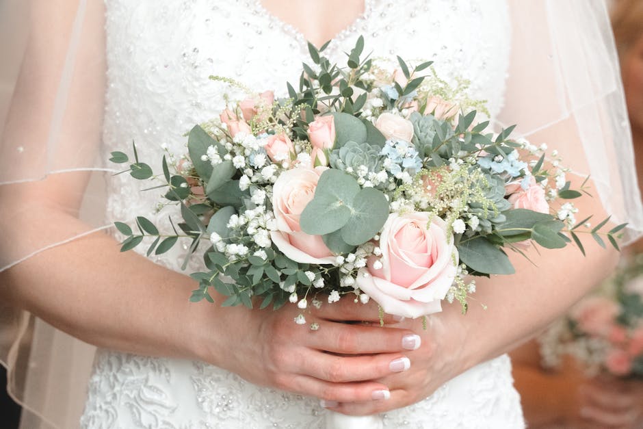 A bride holding a floral bouquet arrangement featuring pale pink roses, white baby's breath, and green eucalyptus leaves. The bouquet is arranged in a round, compact style with a mix of soft pastel co