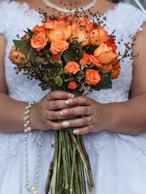 A bride in a white lace wedding dress holds a bouquet of fresh flowers, including orange roses and smaller orange blossoms, with dark green foliage and delicate sprigs of baby's breath. The bouquet is