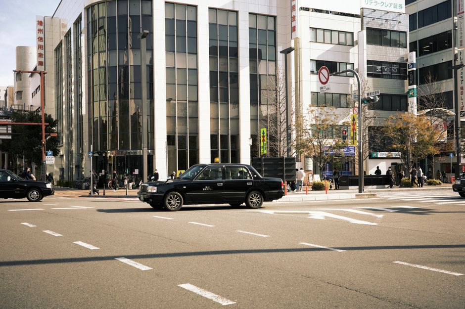 A busy city street scene featuring a modern glass-fronted office building with a white façade, surrounded by tall structures with various signs and advertisements. In the foreground, a black taxi cab
