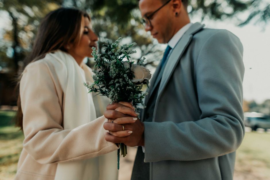 A person in a beige coat holding a bouquet of fresh flowers, including white roses and green foliage, with textured petals and vibrant leaves. The bouquet is wrapped in light green paper and tied with